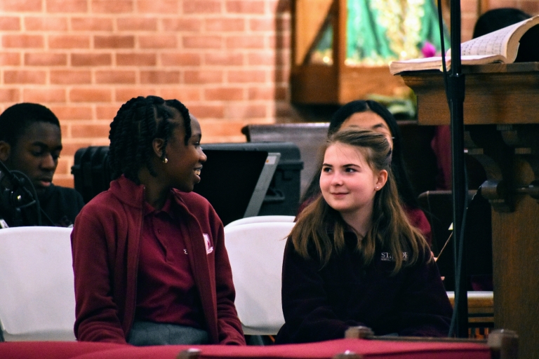 2 6th grade girls smile at one another while singing in the choir during Mass